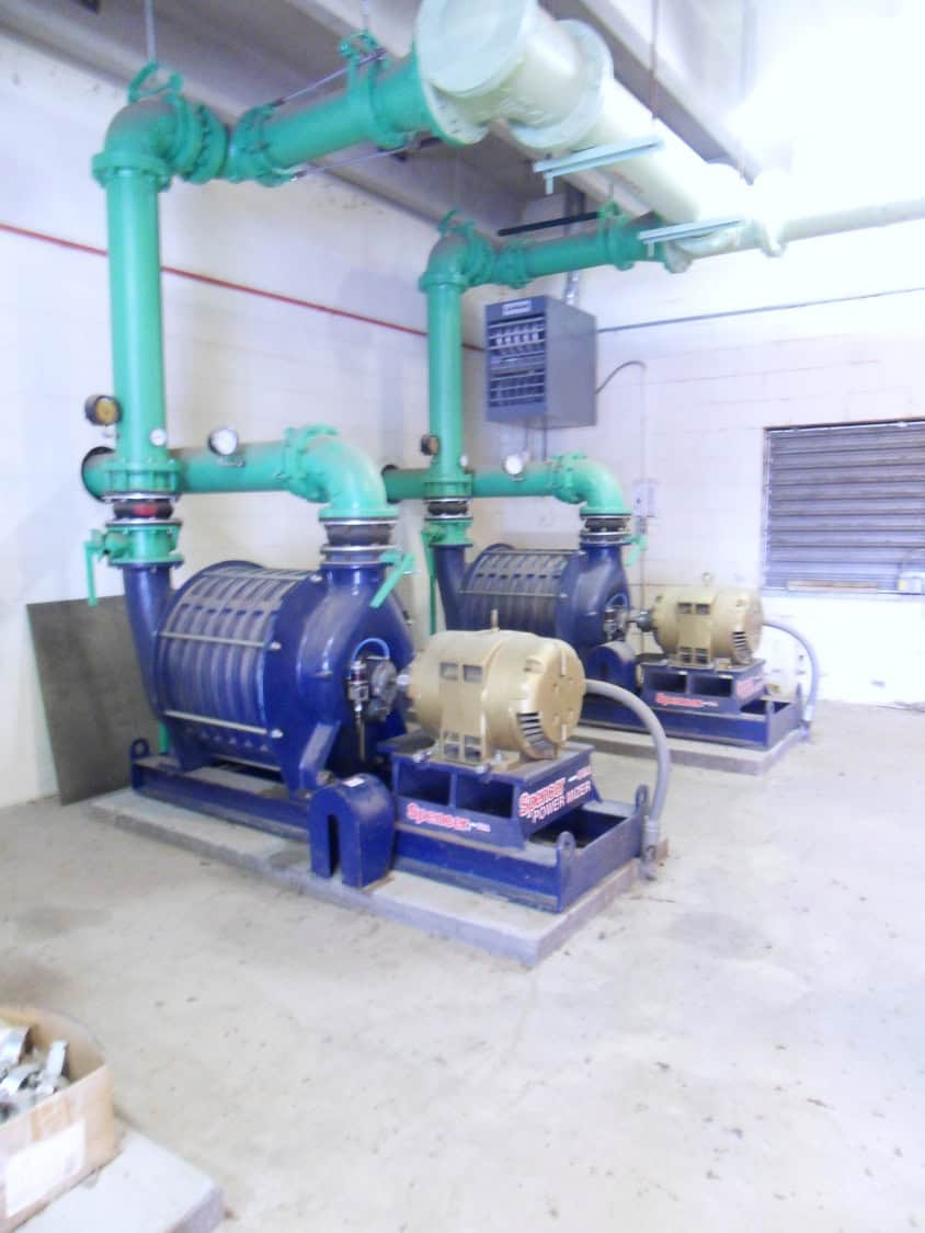 Two large blue industrial pumps driven by beige electric motors, connected to green overhead pipes; they sit on concrete pads inside a white cinderblock utility/boiler room with an electrical panel.

Text in image: "Spencer" and "POWER BLOWER" (on machine bases)