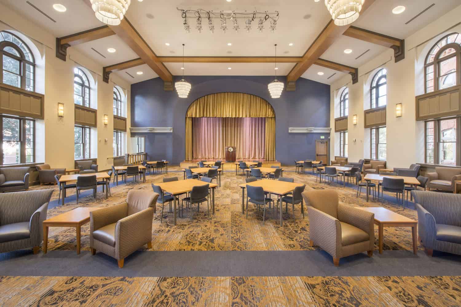 Stage with golden curtain sits at room's center, flanked by a podium; multiple tables and chairs are arranged for small groups across a high‑ceilinged hall with arched windows and chandeliers.