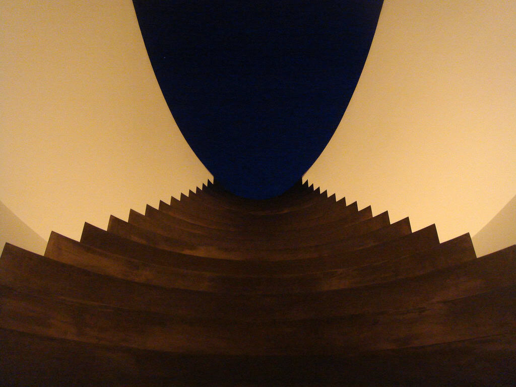 A curved, dark wooden staircase ascends in stepped zigzag tiers toward an oval deep-blue opening, flanked by smooth warm cream walls, photographed from below.
