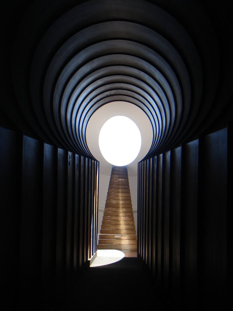 A concentric, ribbed tunnel frames a rising wooden staircase leading toward a bright circular aperture, light casting a semicircular patch on the dark passage's floor.