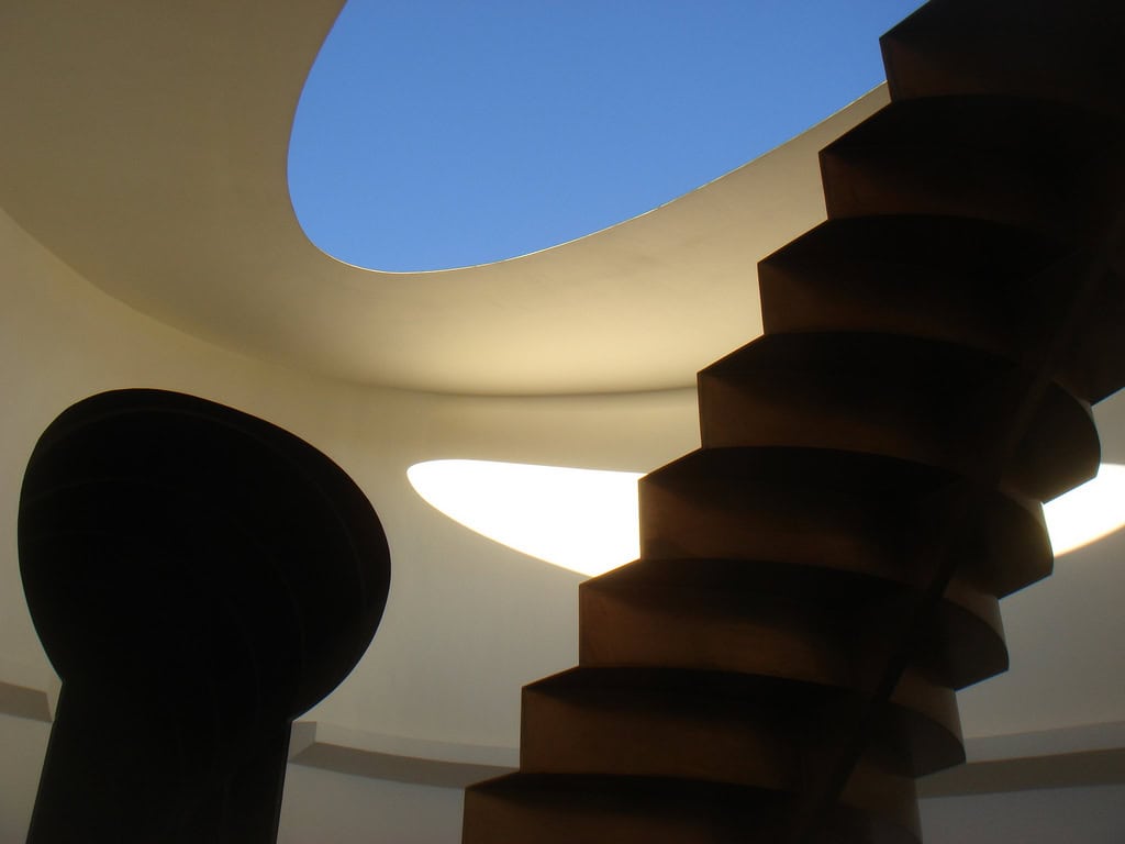 A dark, sculptural spiral staircase curves upward toward an oval skylight, casting layered shadows inside a white, circular atrium where bright blue sky is visible through the opening.