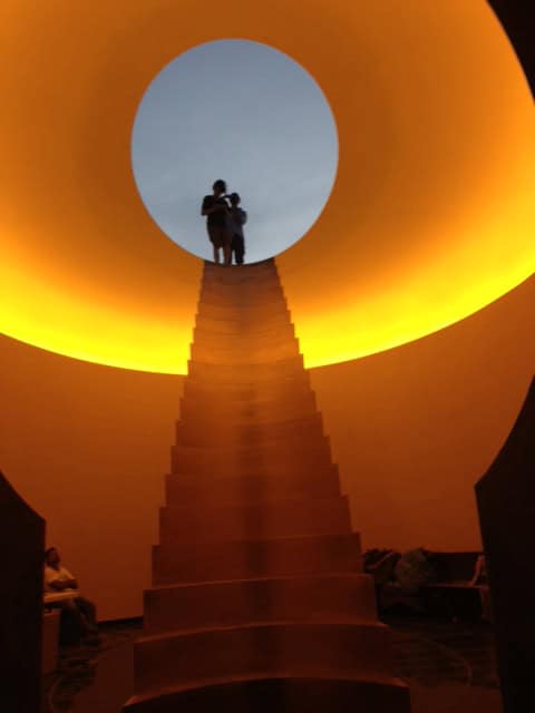 Steep, central staircase ascends toward a circular skylight where two silhouetted figures stand; warm yellow ring of light lines the dome interior, dim seating and people visible at lower sides.