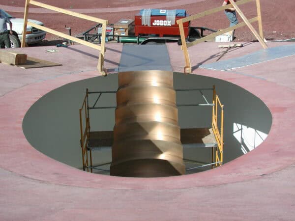 Bronze spiral column descends through a circular rooftop opening while scaffolding surrounds it; wooden guardrails and tools lie on the pink roof near a red toolbox labeled "JOBOX".