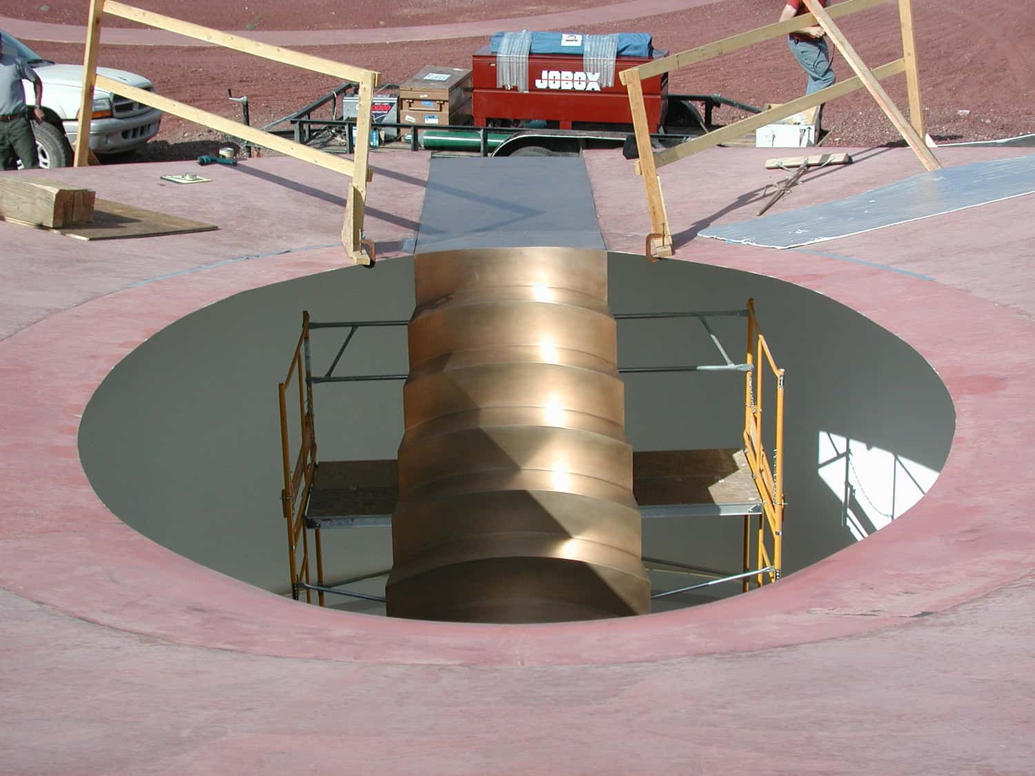 Bronze spiral column descends through a circular rooftop opening while scaffolding surrounds it; wooden guardrails and tools lie on the pink roof near a red toolbox labeled "JOBOX".