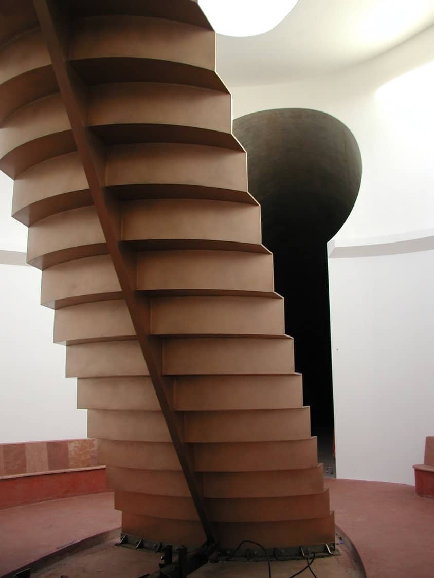 A wide, brown, spiraling staircase twists upward within a white, circular gallery space beneath a skylight, a dark rounded opening visible behind it.