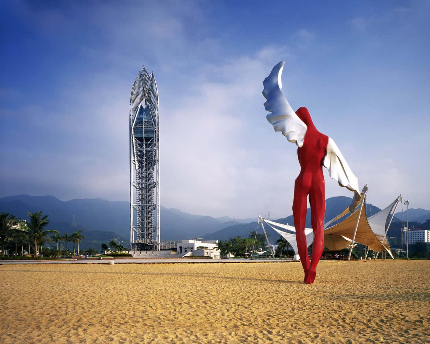 Red winged human sculpture stands with an arm and wing raised on a sandy plaza, framed by a tall latticed tower, white canopies, palm trees and misty mountains.