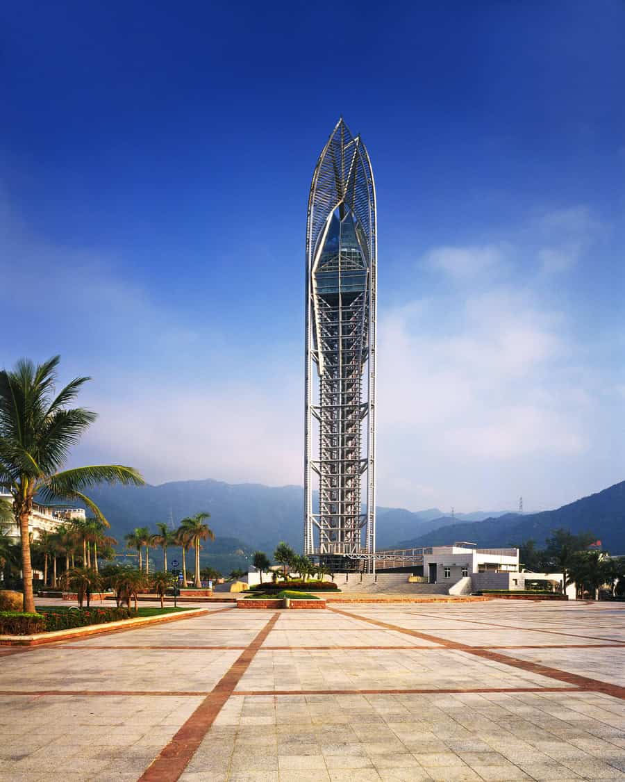 A slender, leaf-shaped skyscraper rises from a tiled plaza, framed by palm trees, with low white buildings and distant green mountains under a clear blue sky.