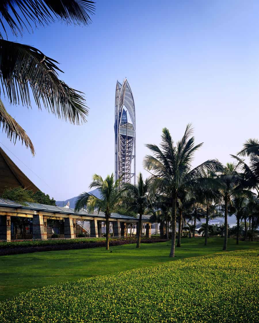 A slender futuristic tower rises above a tropical park, framed by palm trees; a covered walkway and manicured lawn dotted with yellow flowers fill the foreground beneath a blue sky.