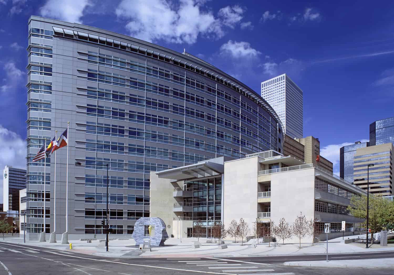 A curved glass-and-steel office building dominates a downtown block, flanked by flagpoles and a stone sculpture on a paved plaza, set against a clear blue sky and surrounding skyscrapers.