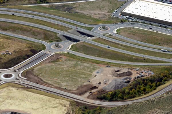 Highway interchange channels cars across twin overpasses and three roundabouts, while earthmoving equipment works on a construction site in surrounding grassy fields beside a large warehouse.