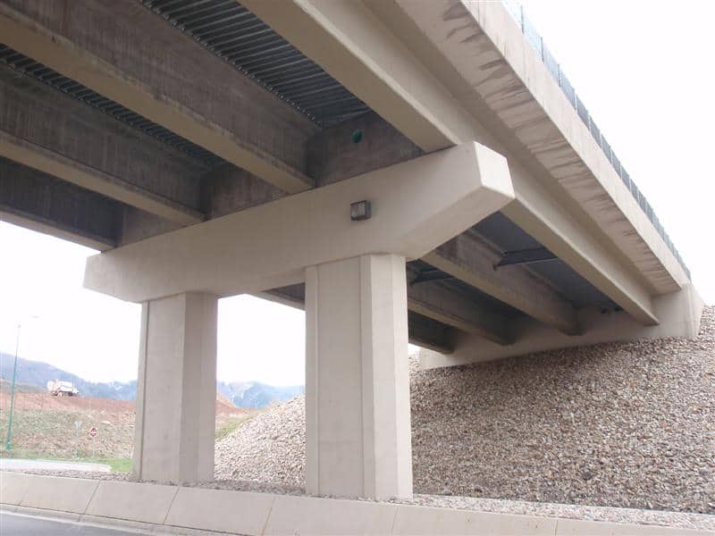 Concrete highway overpass supported by two wide pillars, spanning over a roadway; girders visible underneath, with a gravel embankment to one side and distant hills under an overcast sky.