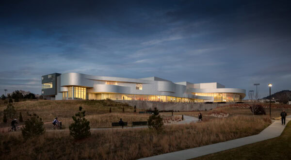 Curved modern building glows from interior lights while people walk and cycle on winding paths through dry grasses at dusk beneath a wide cloudy sky; small unreadable signage on left.