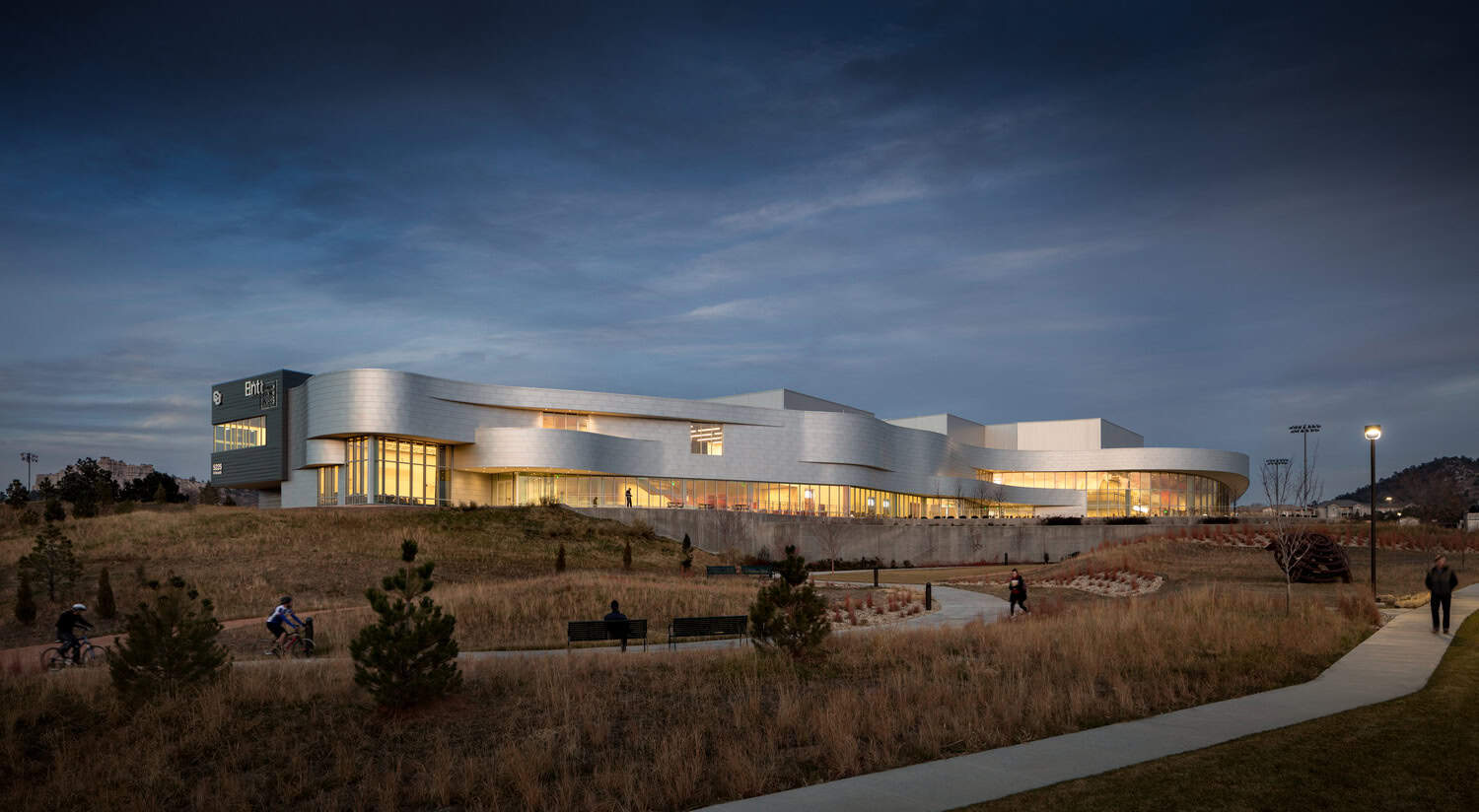 Curved modern building glows from interior lights while people walk and cycle on winding paths through dry grasses at dusk beneath a wide cloudy sky; small unreadable signage on left.