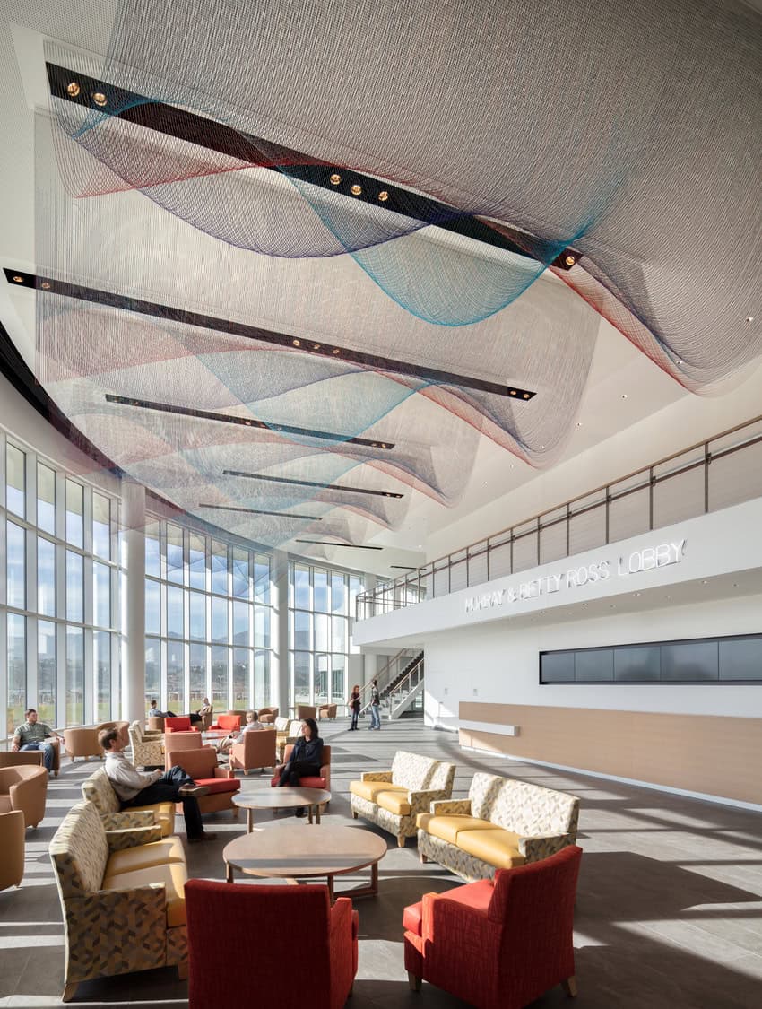 Suspended multicolored mesh sculpture undulates above lounge seating in a spacious, sunlit glass-walled lobby. Text on balcony: "MURRAY & BETTY ROSS LOBBY".