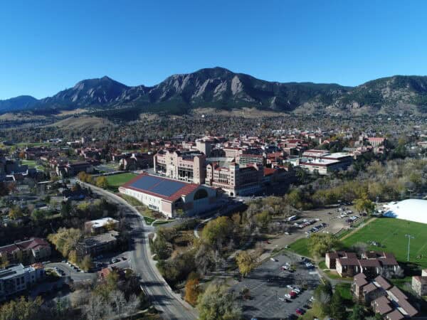 Campus buildings cluster around a large solar-topped arena, stretching across a valley; foothills and rocky Flatirons rise behind, framed by trees and a clear blue sky.