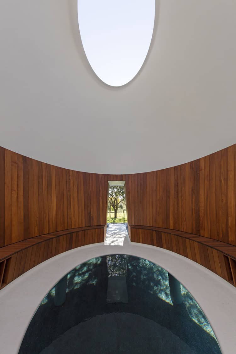 A circular reflecting pool mirrors an oval skylight and trees, framed by curved wooden walls and benches in a domed pavilion with a narrow doorway opening to a sunlit lawn.