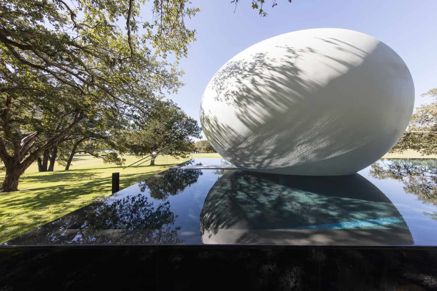 A large smooth white egg-shaped sculpture rests on a glossy black reflecting pool, its shadowed tree patterns mirrored in the water, set in a sunlit grassy park with sprawling oak trees.