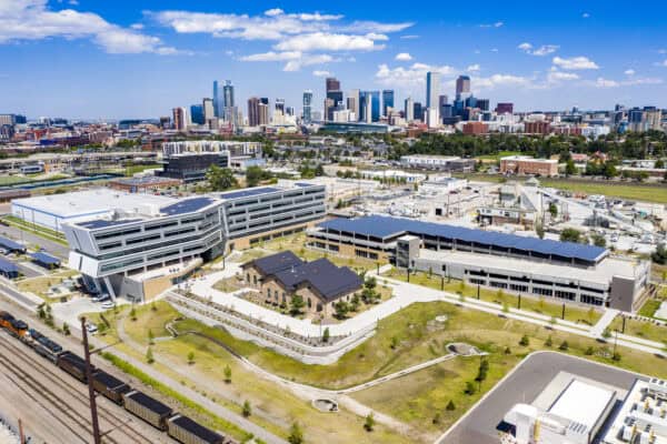A modern office complex with solar-paneled roofs occupies landscaped grounds, adjacent to freight railroad tracks, with a brick historic building in the courtyard and a downtown skyline visible beyond.