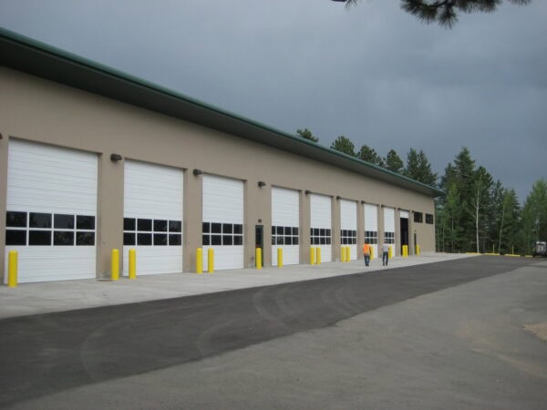 Beige industrial building with multiple closed white roll-up garage doors; two workers walk along its concrete apron past yellow bollards; asphalt driveway and pine trees under gray, overcast sky.