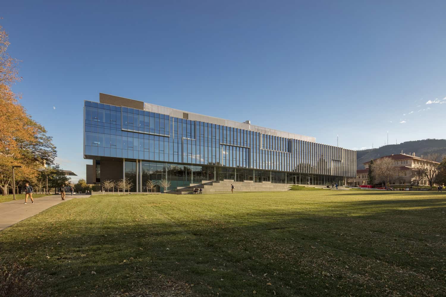 A modern glass-and-steel academic building reflects late sunlight, overlooking a broad grassy quad where students walk and sit, set against a clear blue sky, autumn trees, and distant hills.