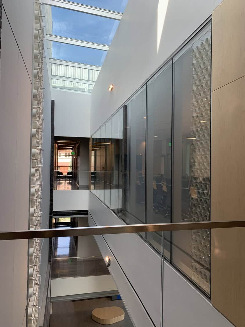 Glass-walled atrium spans three levels under a skylight, connecting office corridors; white decorative wall and glass partitions frame reflective floors, wooden accents, and distant seating at a sunlit end.