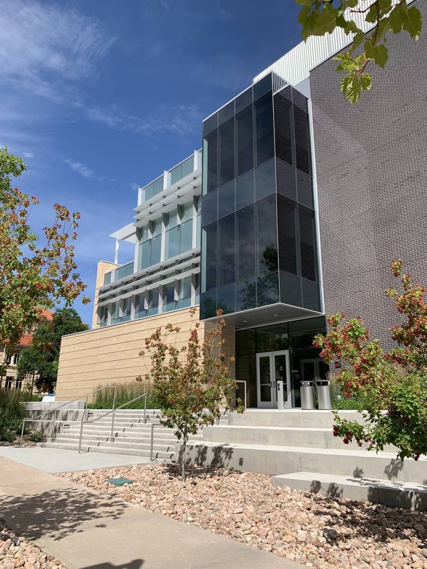 Modern multi-story glass-and-brick building faces the viewer, its entrance set behind concrete steps while small trees and a rocky landscape flank the sidewalk under a clear blue sky.