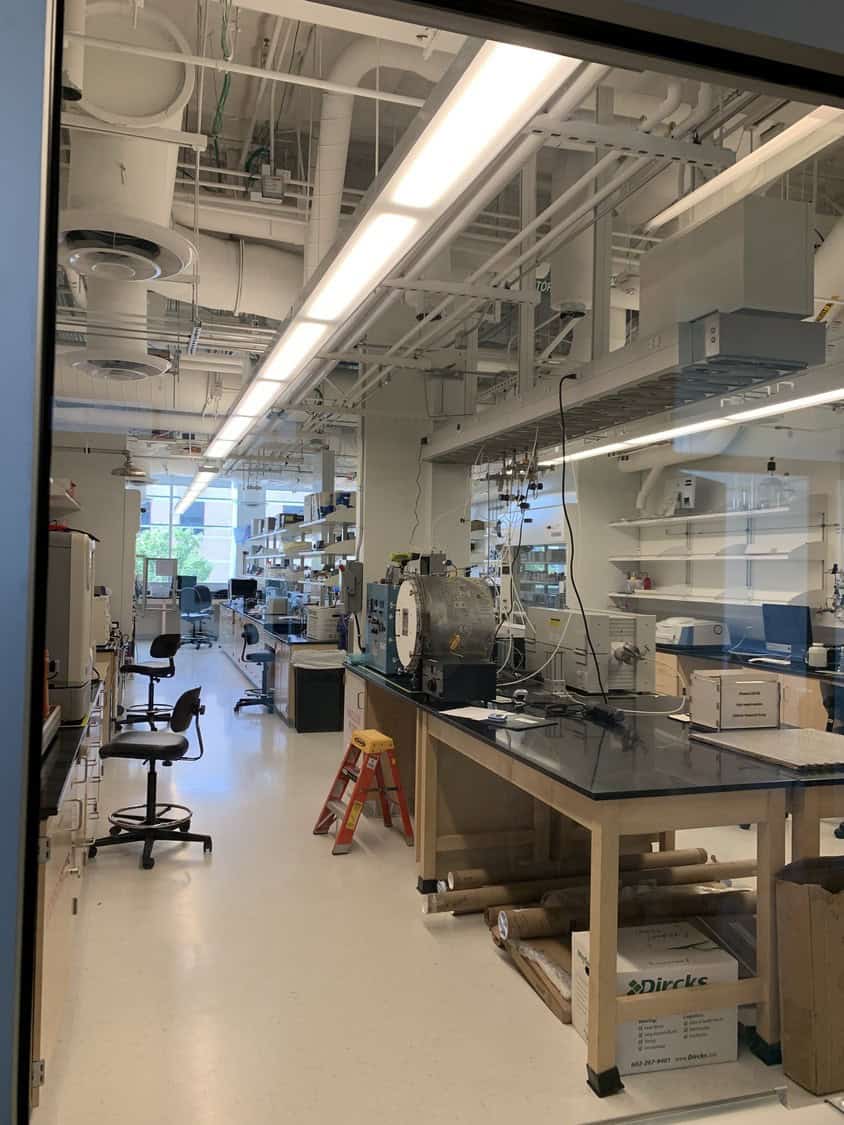 A lab bench supporting scientific instruments and cables, technician stools, and an orange step ladder, arranged along a long, bright research laboratory with overhead ducts, shelving, and windows.

Text found in image (transcribed): "Dircks"