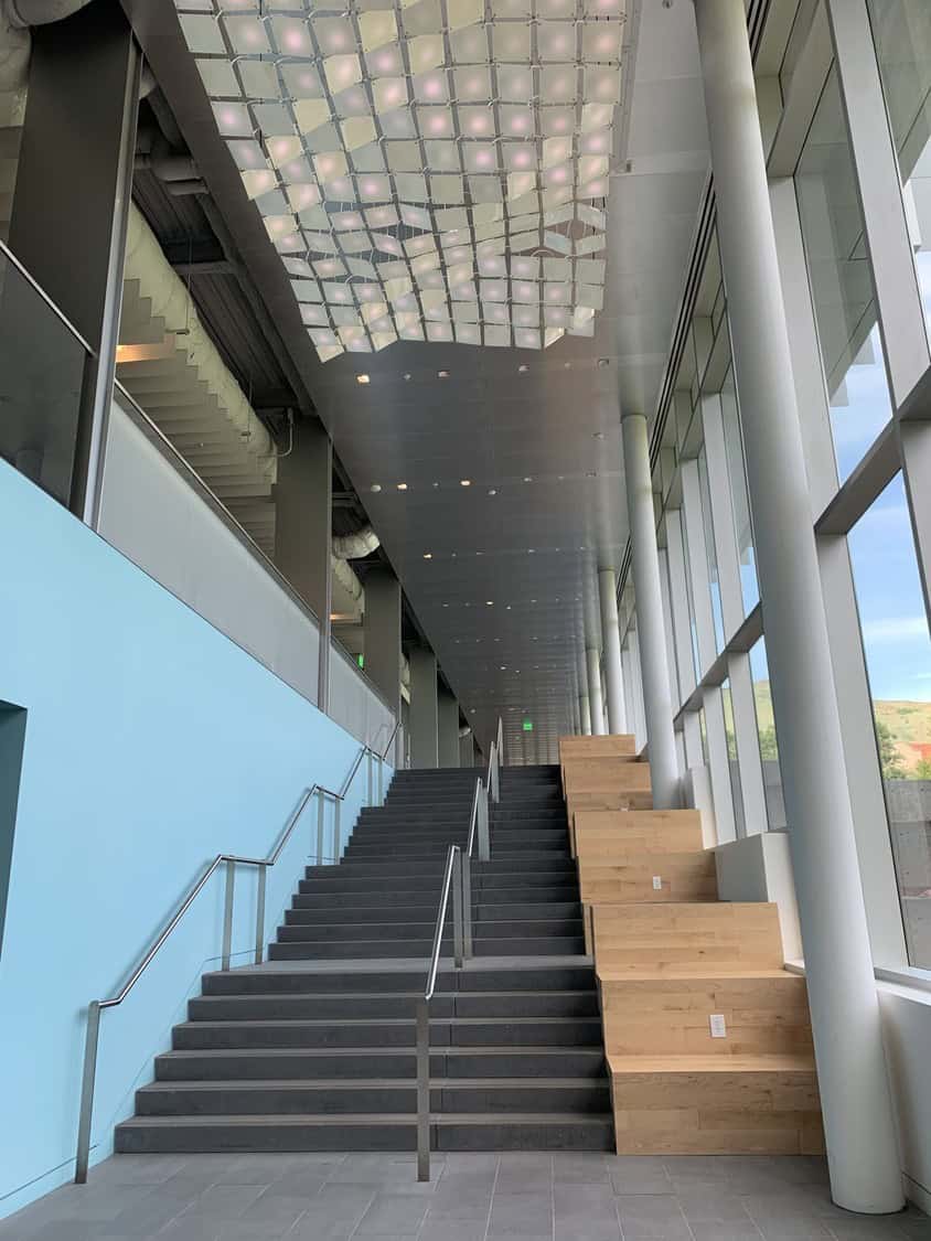 Wide dark staircase rises toward a distant corridor; adjacent wooden stepped benches sit under tall floor-to-ceiling windows, pale blue wall and a patterned illuminated ceiling in a modern atrium.