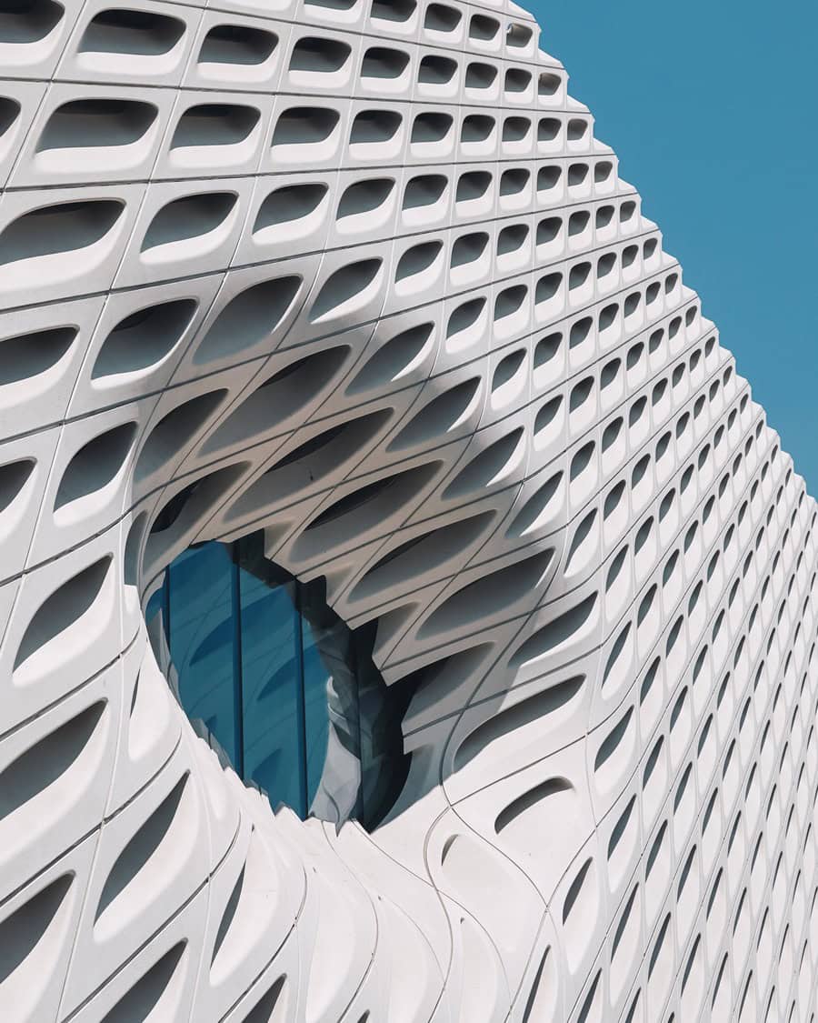 White perforated building façade with repeating teardrop-shaped apertures curves outward, forming an oval opening that exposes blue glass windows, set against a clear, turquoise sky.