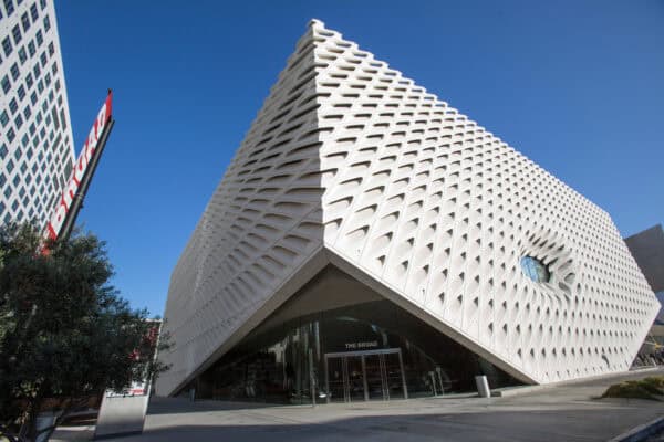 White honeycomb‑façade museum building rises to a corner, revealing a window; sits on a paved plaza under clear blue sky, glass entrance labeled "THE BROAD", red banner "BROAD".