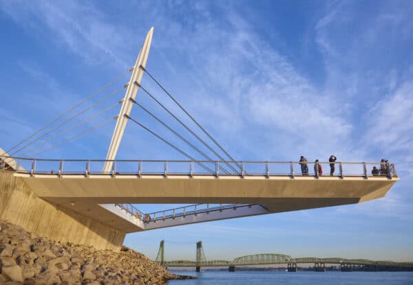 White cable-stayed platform juts over a rocky shore; people stand along its railing gazing across a calm river under a clear blue sky, a green truss bridge visible in the distance.