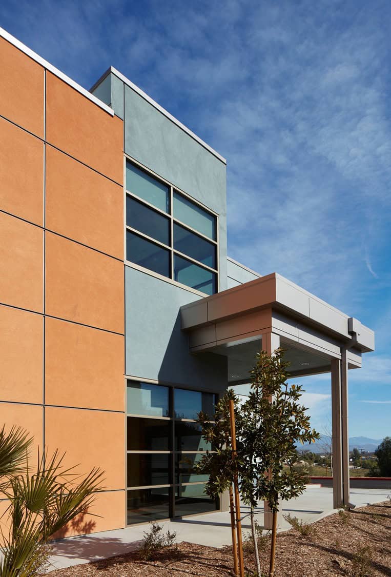 Modern two-story building façade features large grid windows and a projecting entrance canopy, with young landscaped shrubs and mulch at the base, under a clear blue sky.