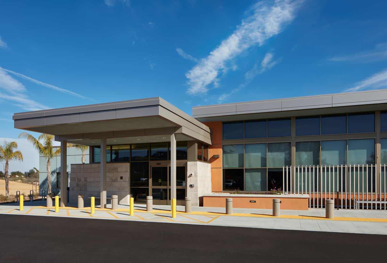 Modern building entrance features a wide flat canopy over automatic glass doors, with yellow bollards and concrete posts protecting the drop-off, set against palm trees and a clear blue sky.