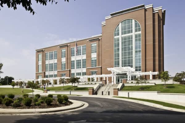 Large red-brick public building dominates a landscaped campus, featuring a tall arched glass façade, an American flag, white pergola and steps leading from a circular driveway under blue sky.