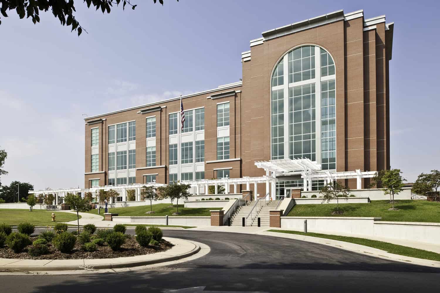 Large red-brick public building dominates a landscaped campus, featuring a tall arched glass façade, an American flag, white pergola and steps leading from a circular driveway under blue sky.