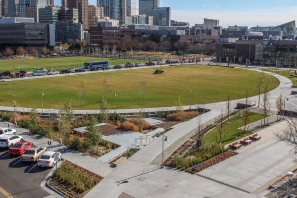 Oval grassy lawn with a riding mower cutting grass, encircled by a curved concrete walkway, landscaped planters, parked cars and mid-rise office buildings in the background.

Visible text: SPEED LIMIT 15; 222‑2222 (on bus); VOTE (small sign).