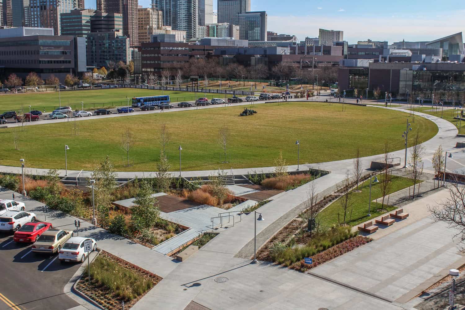 Oval grassy lawn with a riding mower cutting grass, encircled by a curved concrete walkway, landscaped planters, parked cars and mid-rise office buildings in the background.

Visible text: SPEED LIMIT 15; 222‑2222 (on bus); VOTE (small sign).