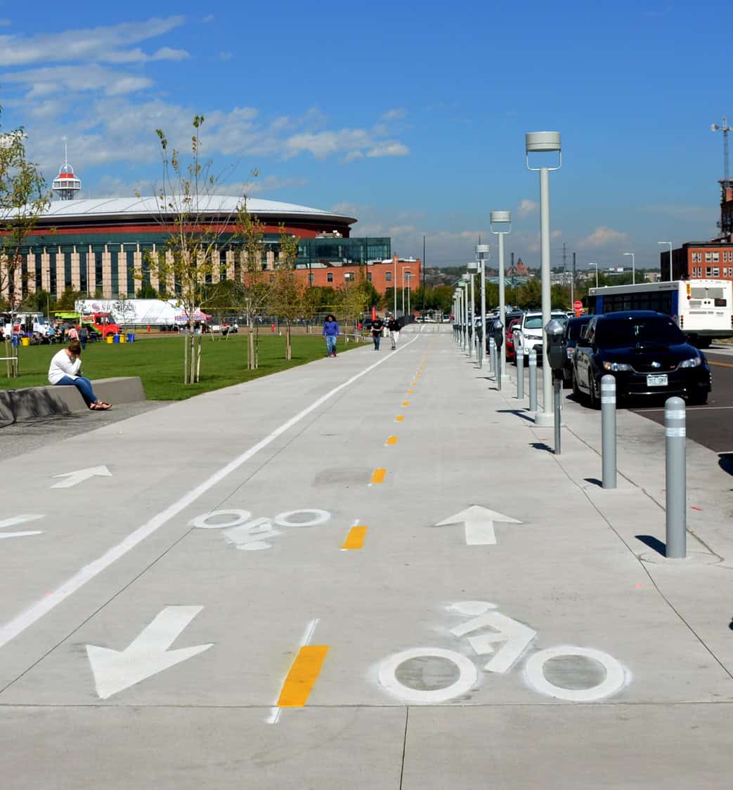 Bike lane painted with arrows and bicycle symbols extends toward stadium; people walk, a person sits in the park beside parked cars and lamp posts under blue sky.