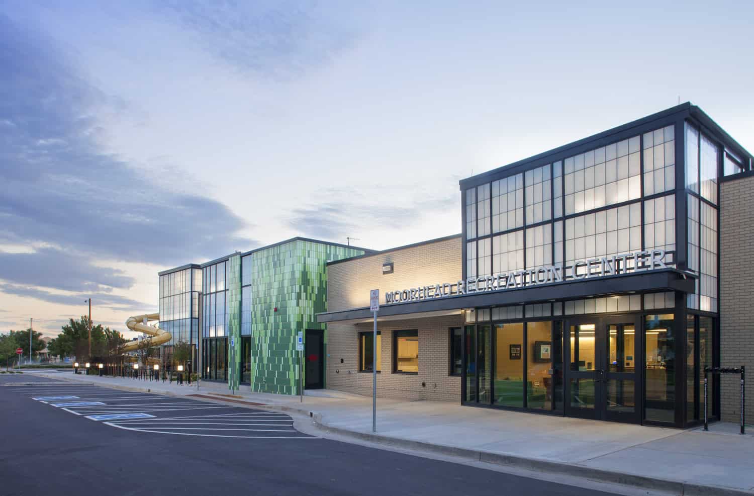 Moorhead Recreation Center building stands at dusk, its glass-and-green-tiled façade illuminated, main entrance lit, empty parking in foreground and a yellow outdoor slide visible to the left.

Text: "MOORHEAD RECREATION CENTER"