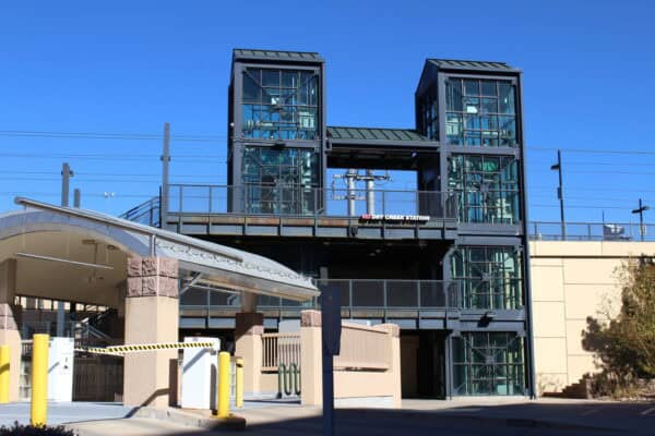 Paired glass elevator towers connect an elevated rail platform above a concrete entrance canopy at a suburban station under a clear blue sky. Text in image: "RTD DRY CREEK STATION".