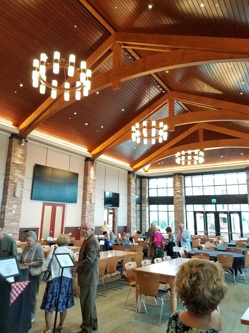 Vaulted wooden ceiling with exposed beams and circular chandeliers hangs above a banquet hall where older adults gather, standing and sitting at long tables near stone columns and large windows.
