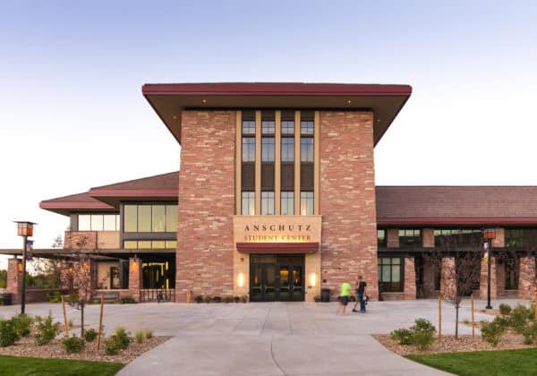 Brick student center tower faces a wide paved plaza; pedestrians pass the glass doors beneath tall windows and a broad overhanging roof at dusk. Text: "ANSCHUTZ STUDENT CENTER".