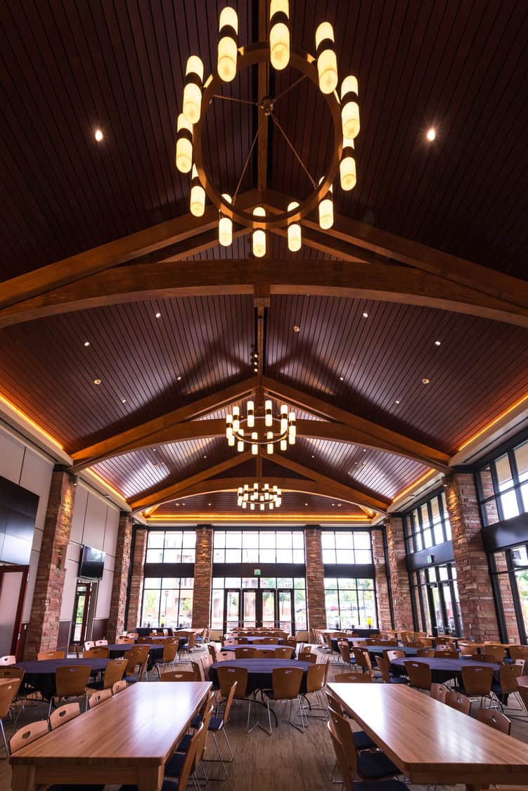 Vaulted wooden ceiling with circular chandeliers hanging above rows of set tables and chairs, inside a spacious dining/event hall framed by stone columns and large glass windows and entrance.