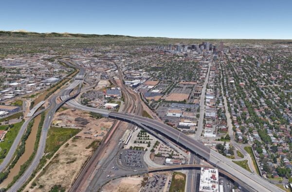 Highway interchange curves and bridges over rail yards and brown river, running through industrial lots and parking fields; residential grid and tree-lined streets extend toward downtown skyline and mountains.