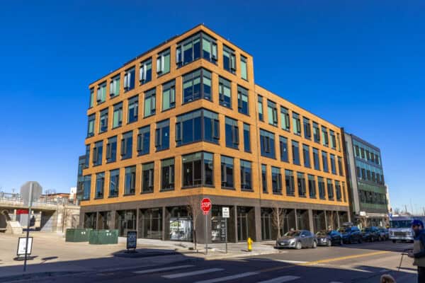Brick five-story office building occupies a street corner, its grid of windows reflecting clear blue sky; cars line the curb and a pedestrian walks a dog near a stop sign.

Transcribed text: STOP; ALL WAY; NO MOTOR VEHICLES.