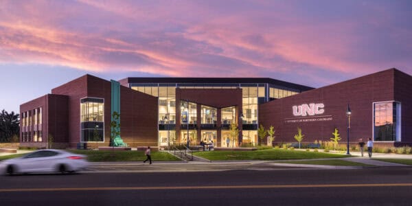 A modern brick university building displays glowing signage reading "UNC" and "UNIVERSITY OF NORTHERN COLORADO"; its glass entrance is lit as people walk and a car passes on a street at dusk beneath pink clouds.