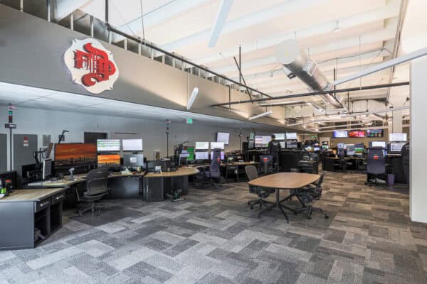Open operations control room: operators monitor multiple screens at curved desks beneath exposed ducts and bright ceiling, in a spacious carpeted facility.

Text visible: "EXIT"; "FIRE 2".
