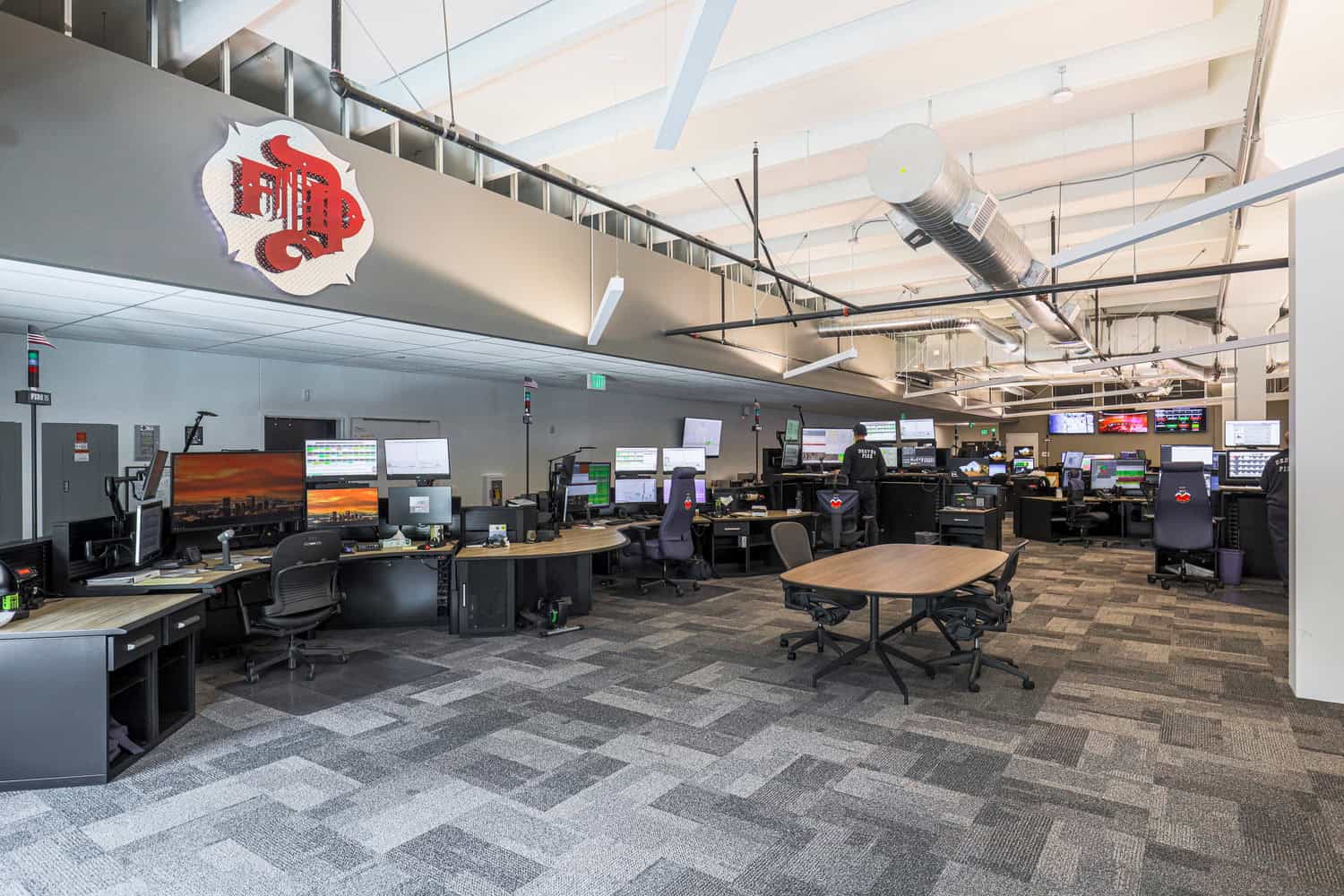 Open operations control room: operators monitor multiple screens at curved desks beneath exposed ducts and bright ceiling, in a spacious carpeted facility.

Text visible: "EXIT"; "FIRE 2".