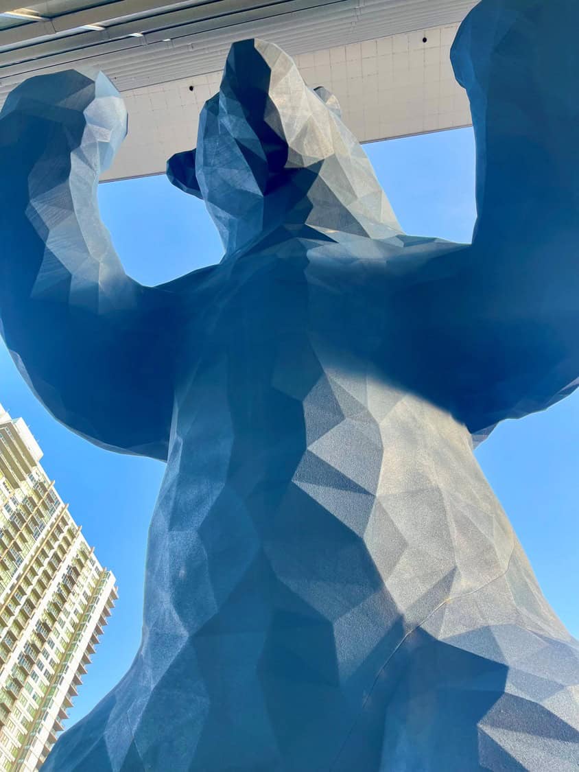A massive faceted blue bear sculpture rears with forepaws pressed against an overhead concourse, beneath a clear blue sky beside a tall apartment building.