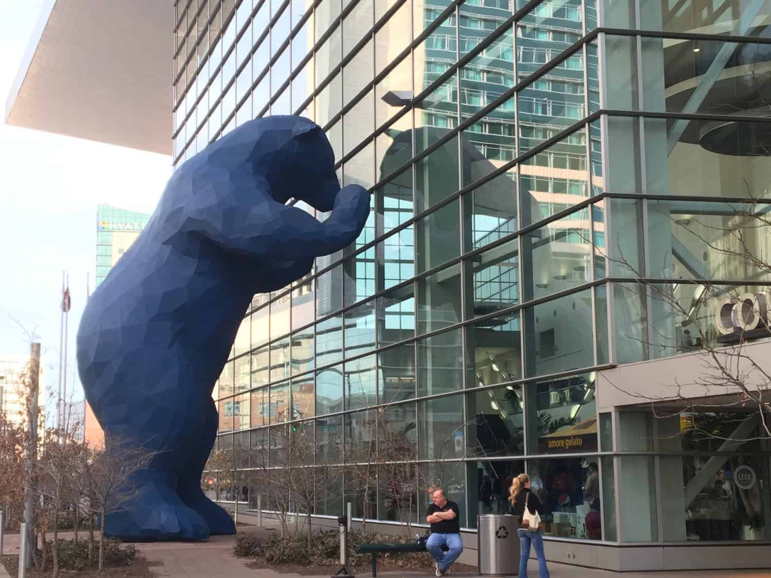 Large faceted blue bear sculpture presses its paws and forehead against a glass building facade in an urban plaza with pedestrians and planters.

Text visible: amore gelato; HYATT; CO; LEED